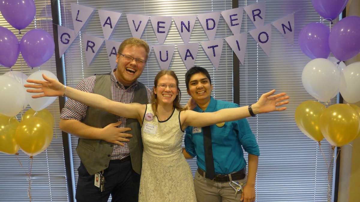 Two males and one female graduates in front of a glass wall with a banner that reads "Lavender Graduation banner"