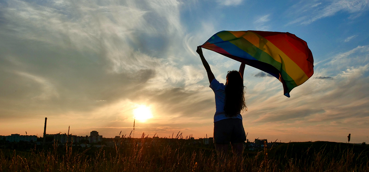 Silhouette of a person holding a PRIDE flag in front of a sunset.
