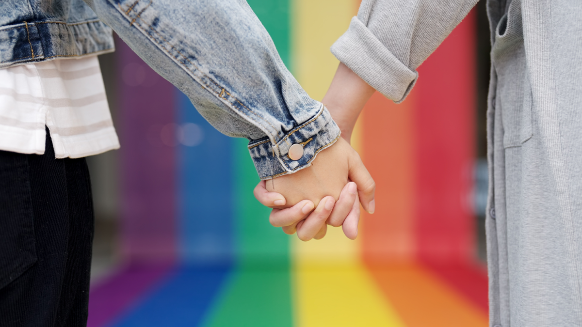 Two gals holding hands in front of a rainbow flag.