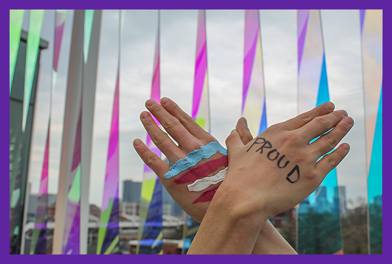 Hands forming a butterfly in front of "Watermak" installation at Crosland Tower. One "wing" says "Proud" and the other one has the trans flag painted on it.