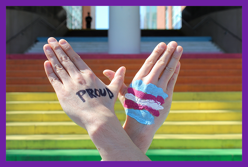 Two hands forming a butterfly. On one "wing" the word "Proud" is written, and on the other one the trans flag has been painted.